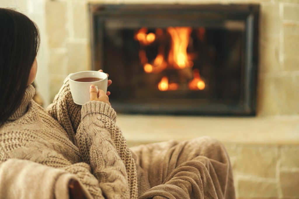 Woman in cozy sweater holding cup of warm tea at fireplace. Heating house with wood burning stove